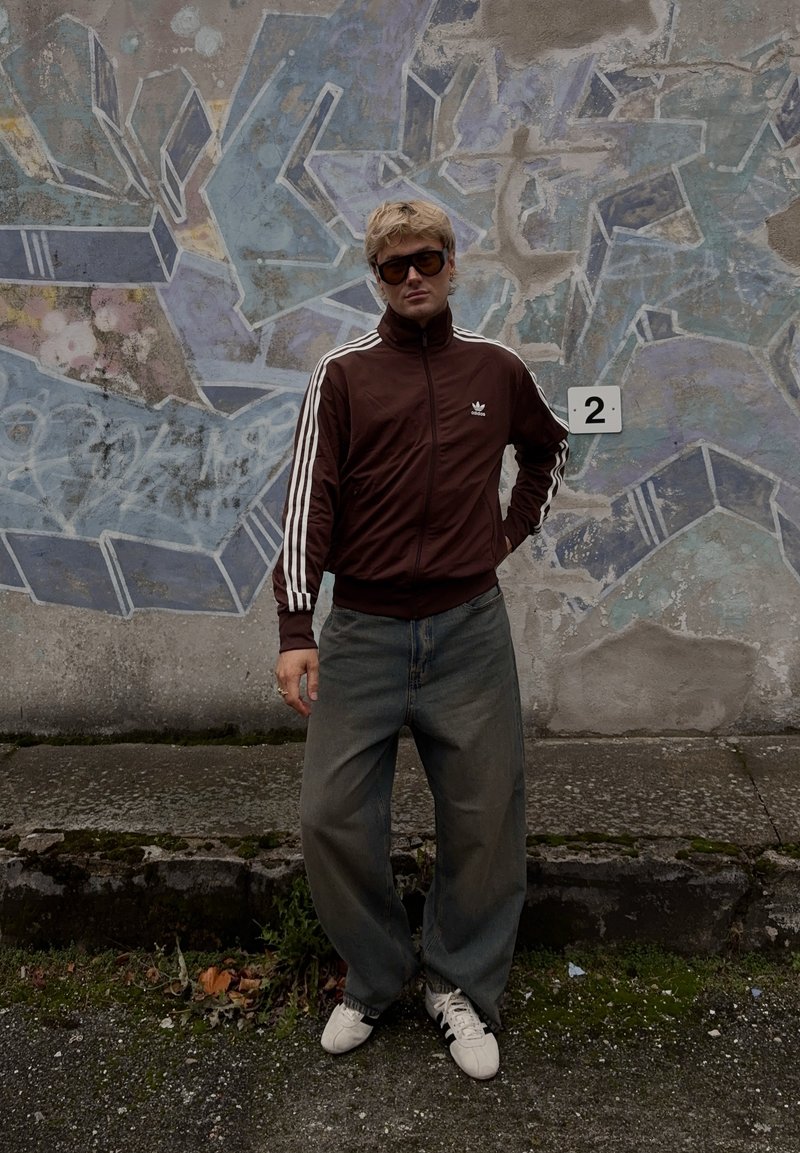 Bruine zip-up sportjack met witte strepen, ruime grijze spijkerbroek en witte sneakers. De achtergrond toont een kleurrijk abstract muurschildering.