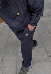Chaqueta rompevientos negra con un pequeño logo amarillo y pantalones de chándal negros con un logo blanco de Nike, combinados con botas grises de cordones sobre pavimento de baldosas.