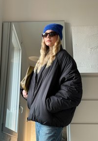 Casaco puffer preto com silhueta oversized, tecido texturizado, fecho com zíper, gorro de malha azul e óculos de sol escuros, segurando uma mala acolchoada verde.