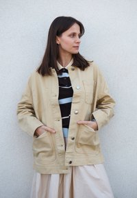 Giacca in tela beige con chiusura a bottoni, due tasche frontali e una vestibilità rilassata. Indossata sopra una camicia a righe e una gonna plissettata.