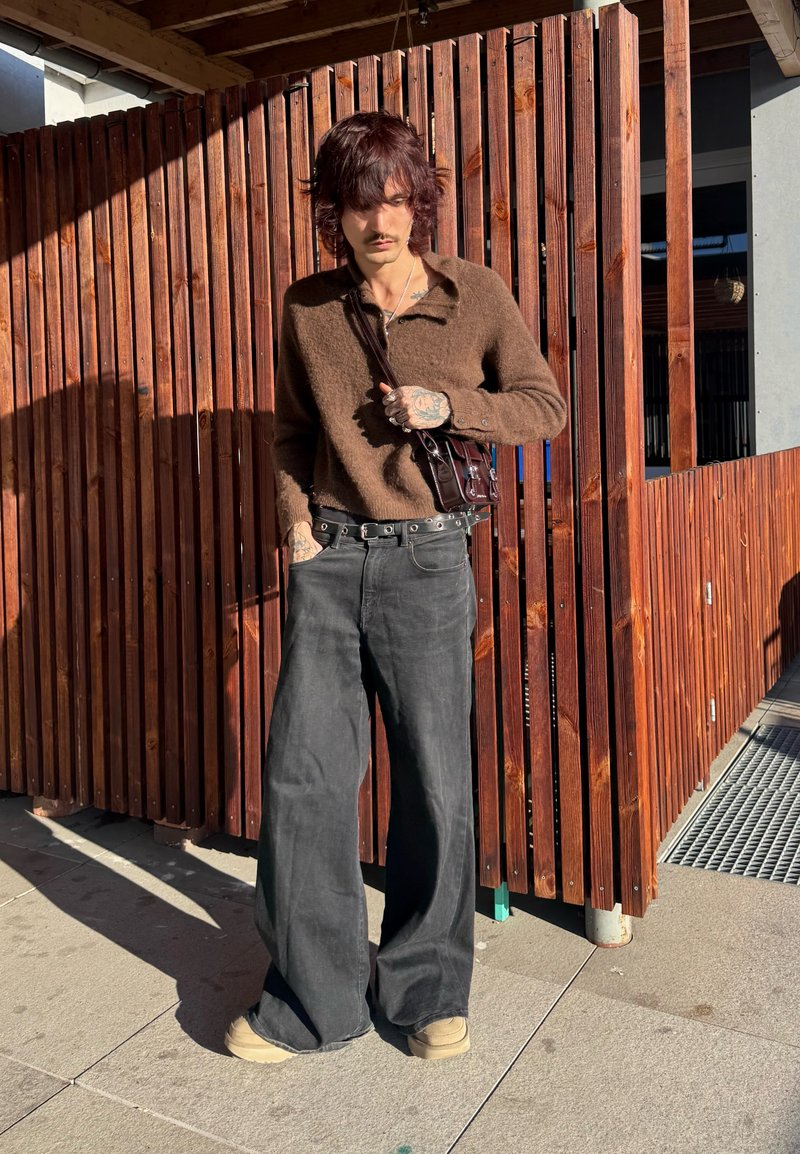 Persona con maglione marrone peloso, pantaloni neri larghi, scarpe beige e una piccola borsa a tracolla scura, in piedi contro una recinzione di legno verticale al sole.