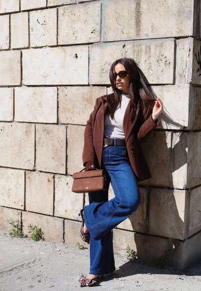 Mujer con gafas de sol, que lleva una blazer marrón, top blanco, vaqueros azules y bailarinas con estampado de leopardo, está de pie junto a una pared de piedra sosteniendo un bolso marrón.
