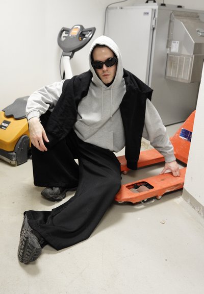 Persona con sudadera gris y chaleco negro, agachada al lado de un transpaleta naranja sobre concreto. Pantalones amplios negros y zapatillas deportivas visibles.