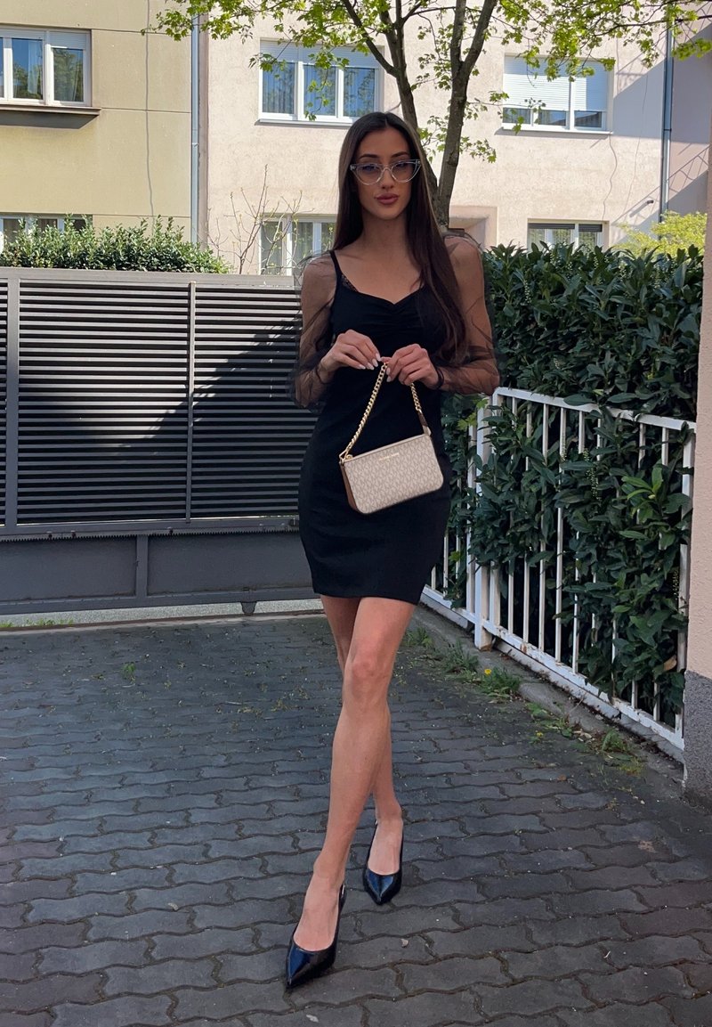 Vrouw met lang donker haar, met een zwarte jurk en hakken, houdt een beige handtas vast en staat op een geplaveid pad bij een woonomheining.