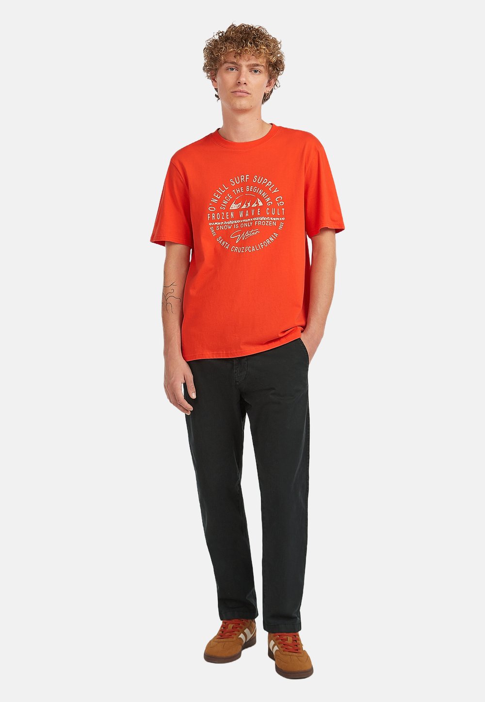 Vermilion katoen t-shirt met een ronde grafische print in het wit, gecombineerd met zwarte broeken en een paar bruine sneakers. Het model staat tegen een witte achtergrond.