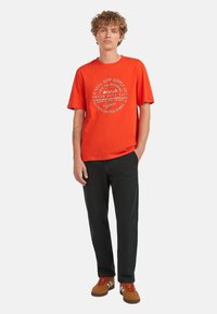 Vermilion katoen t-shirt met een ronde grafische print in het wit, gecombineerd met zwarte broeken en een paar bruine sneakers. Het model staat tegen een witte achtergrond.