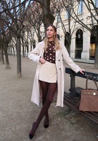 Femme en manteau beige, top à pois marron, jupe crème et collants marron, appuyée sur un banc avec un grand sac à main marron dans un parc bordé d'arbres sans feuilles.