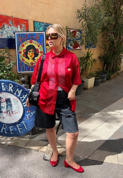 Camiseta de malla roja de Nike, pantalones cortos de mezclilla negros, zapatos rojos y una pequeña bolsa negra con tachuelas. Enmarcado por un colorido arte urbano en un entorno vibrante.