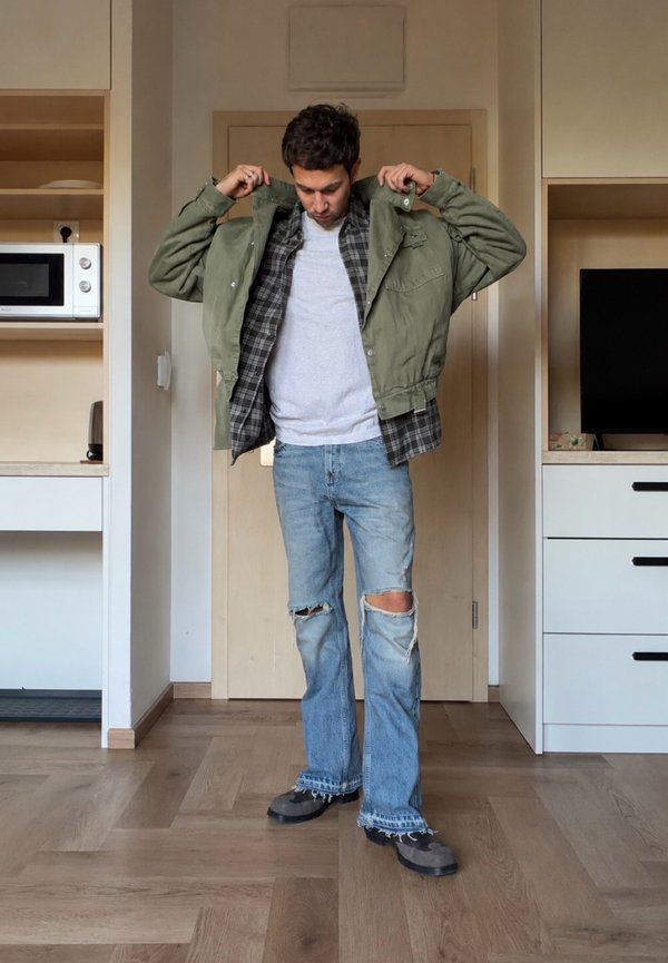 Hombre con vaqueros rotos y camisa gris poniéndose una chaqueta verde en una habitación con suelo de madera, microondas y mueble de televisión.