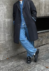 Μαύρο μάλλινο παλτό πάνω από ένα μπλε τζιν πουκάμισο, ανοιχτό μπλε παντελόνι καμπάνα και μαύρα παπούτσια. Πόζαρε μπροστά από έναν υφασμένο τσιμεντένιο τ οίχο.