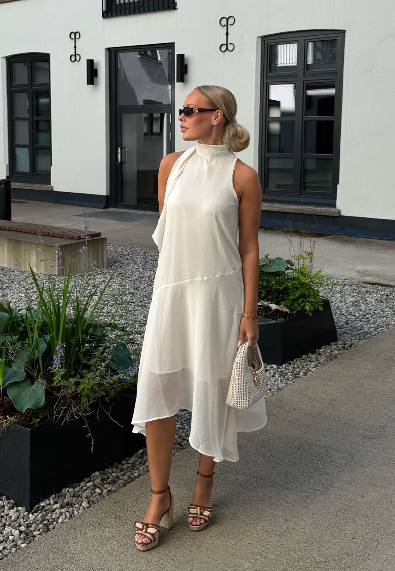 Femme en robe blanche sans manches, sandales à talons marron clair, lunettes de soleil et sac à main blanc, debout sur un chemin en pierre devant un bâtiment blanc.