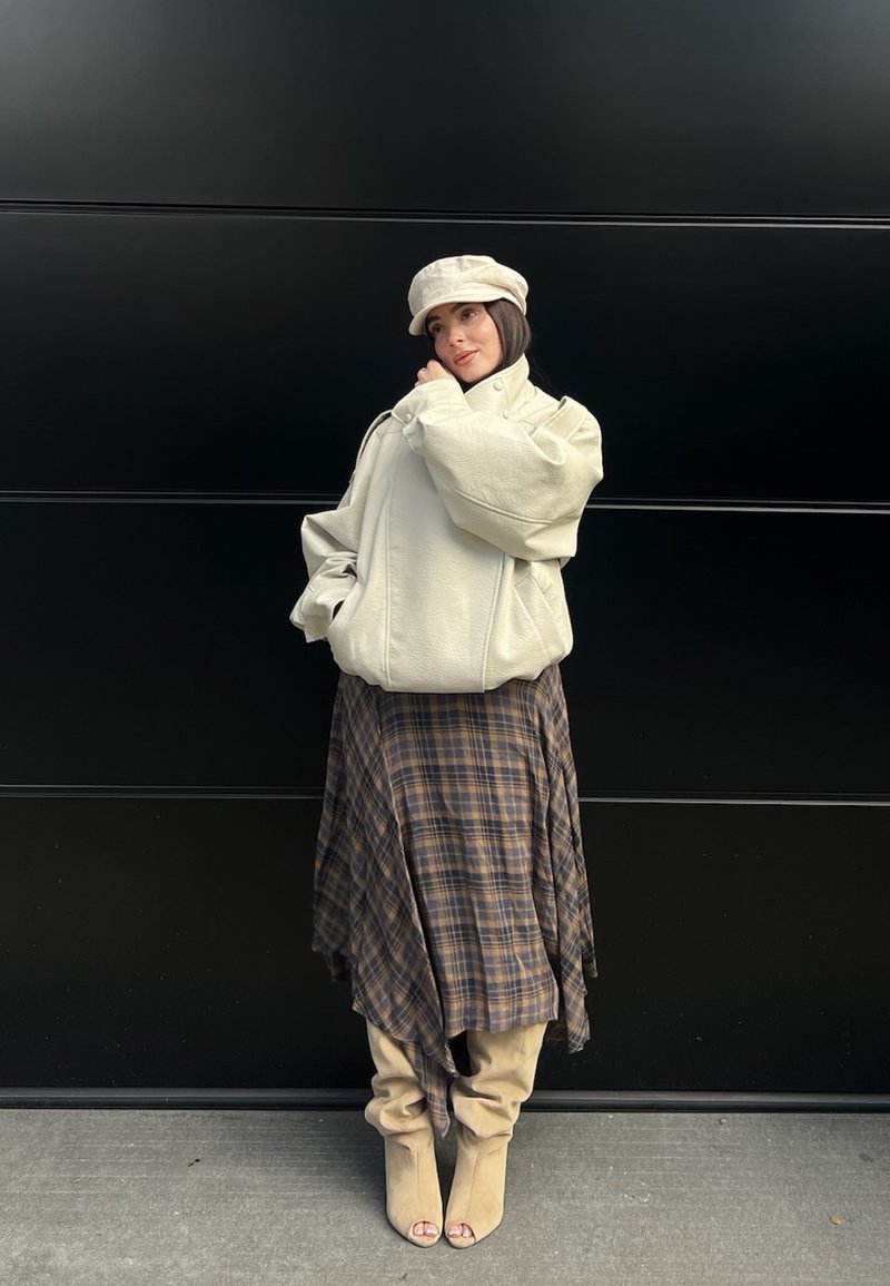 Mujer posando contra pared negra, con gorra crema y chaqueta de gran tamaño, falda asimétrica de cuadros y botines beige con punta abierta.