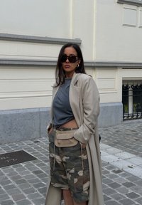 Beige lange jas, grijze T-shirt en camouflage cargoshorts met een tan heuptasje. Grote zonnebril en minimale accessoires. Textuur van het wegdek als achtergrond.