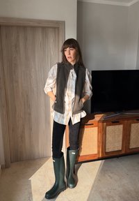 Femme debout à l'intérieur, portant une chemise à carreaux, un gilet sans manches, un pantalon noir et de grandes bottes en caoutchouc vertes, près d'une armoire en bois et d'une télévision.