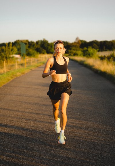 Sujeción deportiva negra y pantalones cortos negros combinados con zapatillas de correr verde claro. Silueta atlética y suave; corredor en un entorno natural al aire libre.