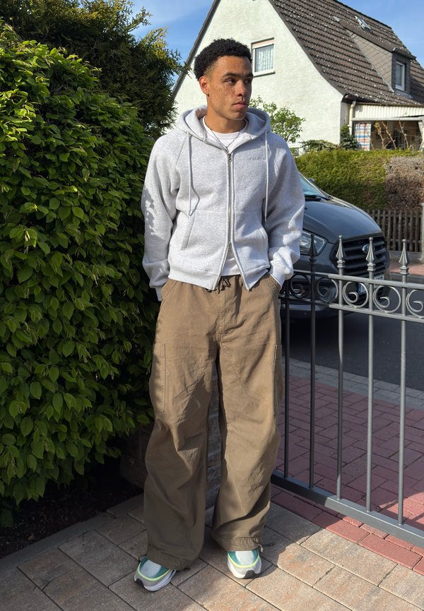 Junger Mann in beigen Hosen, grauem Kapuzenpullover mit Reißverschluss und Turnschuhen, der draußen neben grünen Büschen und einem Metallzaun vor einem Haus und einem geparkten Auto steht.
