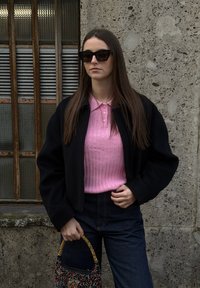 Femme aux cheveux longs bruns portant des lunettes de soleil noires, une veste noire, un polo rose côtelé, un jean foncé, tenant un sac à poignée de perles colorées près d'un mur en béton texturé.