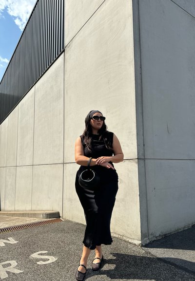 Vestido negro sin mangas, ajustado con un dobladillo en forma de ondas. Bolso tipo cubo negro y zapatos de slip-on. Fondo de concreto, cielo azul claro con nubes.