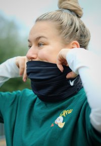 Vrouw met blond haar in een knot die een zwarte Nike nekwarmer aanpast, gekleed in een groene shirt met een kleine kleurrijke grafiek en lange witte mouwen buiten.