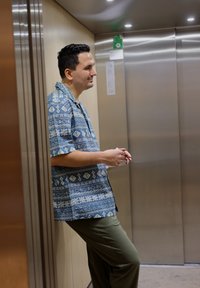 Chemise à manches courtes bleue avec des motifs géométriques blancs ; pantalon vert ; se tient dans un ascenseur aux parois métalliques avec un éclairage au plafond.