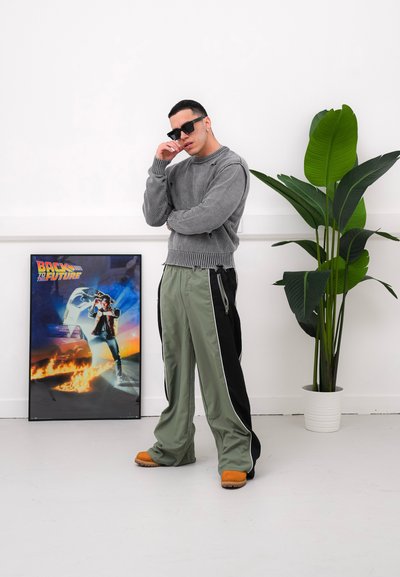 Suéter de punto gris con dobladillo acanalado, pantalones de chándal verde oliva y negros con rayas laterales, zuecos naranjas y gafas de sol negras. Cartel de Volver al Futuro cercano.