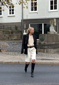 Czarna marynarka, beżowy sweter, białe szorty i czarne kozaki do kolan. Stylizacja charakteryzuje się dopasowanym krojem oraz kontrastami w kolorze i strukturze.