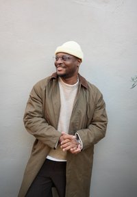 Trenci verde din bumbac cu guler maro, pulover bej și pantaloni în carouri întunecați. Căciulă galbenă și ochelari rotunzi completează ținuta.