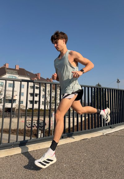 Joven corriendo al aire libre por un camino pavimentado junto a una barandilla de metal, vistiendo una camiseta sin mangas ligera, pantalones cortos y zapatillas blancas para correr bajo un cielo azul despejado.