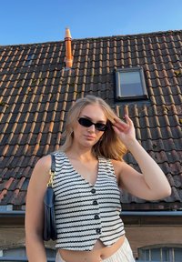 Fehér és fekete csíkos kötött mellény gombokkal, fekete kézitáskával párosítva. A háttérben cserépfedés és kék ég látható.