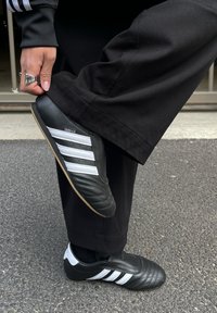 Scarpe sportive nere con strisce bianche, con una tomaia testurizzata e un design a profilo basso. Una persona indossa pantaloni neri larghi mentre solleva un piede.