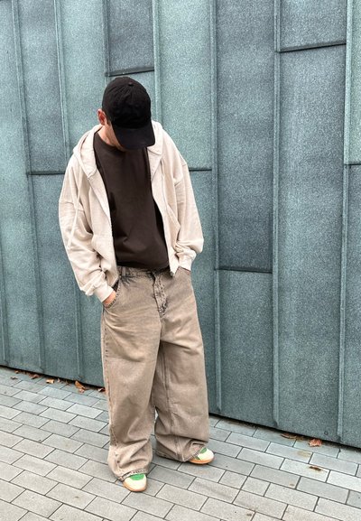 Sudadera oversize de color beige claro, camiseta marrón oscura y pantalones beiges de pierna ancha con un aspecto deslavado. Gorra negra y zapatillas con acentos verdes.