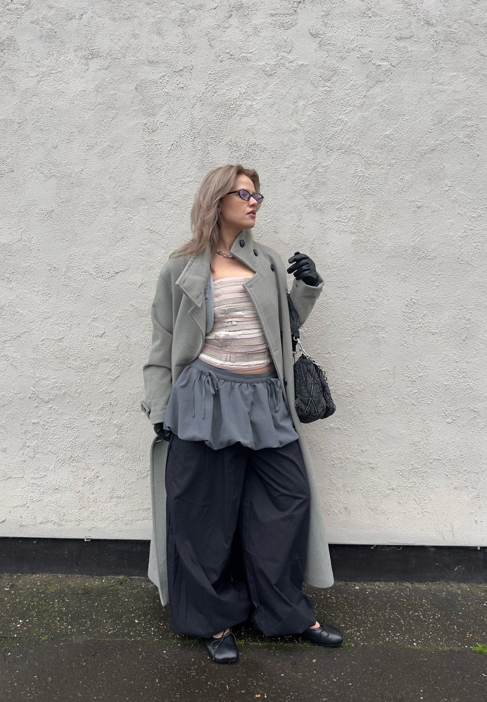 Long manteau gris porté sur un haut à rayures claires, associé à une jupe péplum grise et un pantalon noir à jambes larges. Des accessoires noirs et des gants complètent la tenue.