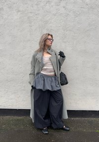 Long manteau gris porté sur un haut à rayures claires, associé à une jupe péplum grise et un pantalon noir à jambes larges. Des accessoires noirs et des gants complètent la tenue.