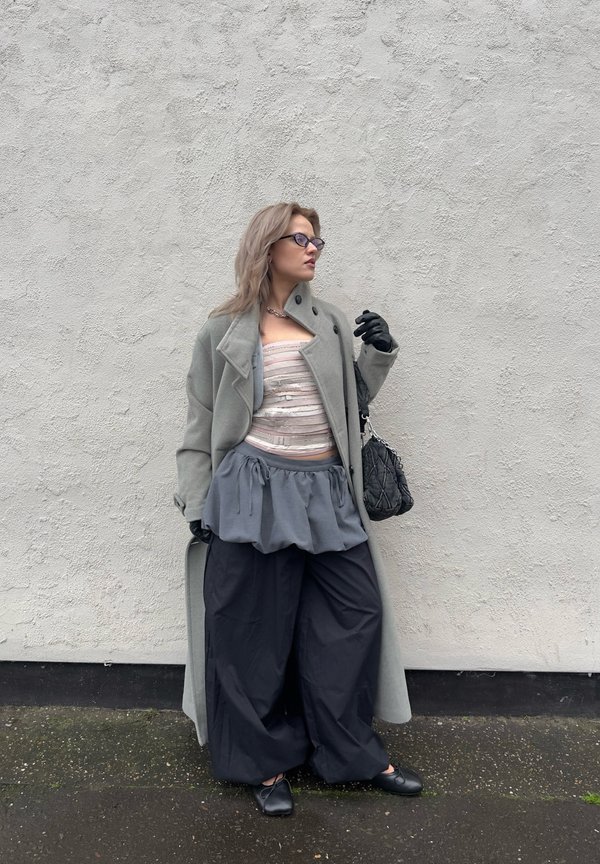 Long grey coat over a light striped top, paired with a grey peplum skirt and black wide-leg trousers. Black accessories and gloves complete the outfit.