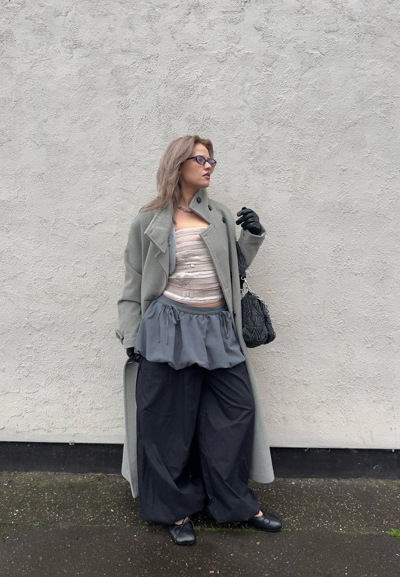 Long manteau gris porté sur un haut à rayures claires, associé à une jupe péplum grise et un pantalon noir à jambes larges. Des accessoires noirs et des gants complètent la tenue.