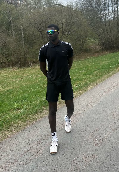 Conjunto deportivo con una camiseta polo negra, pantalones cortos negros y zapatillas blancas. Presenta unas gafas de sol espejadas y coloridas. Césped y árboles de fondo.