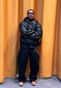 Veste bomber en cuir noir sur une chemise noire, associée à un pantalon noir ample et des chaussures beiges. Les accessoires incluent une chaîne et des lunettes.