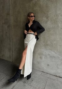 Camisa preta transparente, saia branca com fenda alta, cinto preto com detalhes em dourado e botas pretas de salto até o tornozelo. Fundo de concreto cinza.