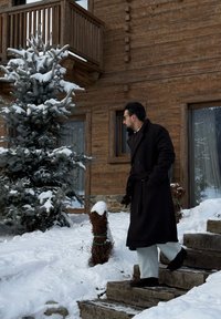 Manteau noir à double boutonnage, associé à un pantalon bleu clair et des bottes foncées, debout sur des escaliers en bois à côté d'un arbre enneigé.