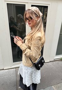 Žena s dlouhými blond vlasy, béžovou bundou, puntíkovaným šátkem na hlavě, slunečními brýlemi a černou koženou taškou používá chytrý telefon na městském chodníku.