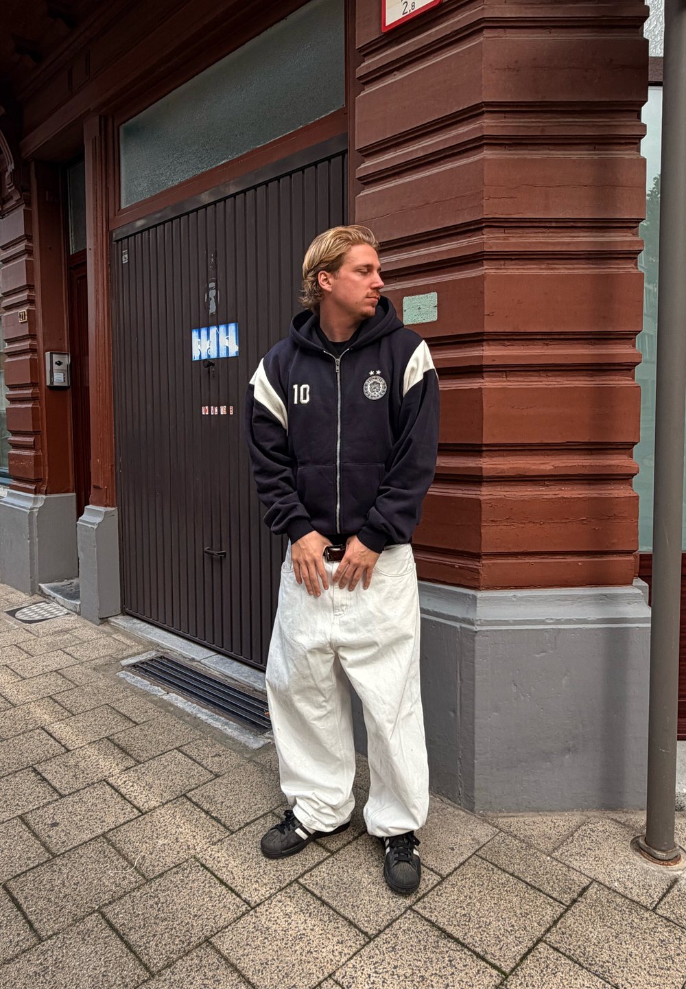 Joven vestido con sudadera negra y pantalones anchos blancos está de pie en la acera junto a la esquina de un edificio de ladrillo rojo, mirando hacia su izquierda.