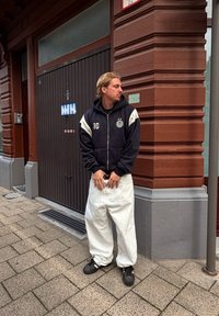 Joven vestido con sudadera negra y pantalones anchos blancos está de pie en la acera junto a la esquina de un edificio de ladrillo rojo, mirando hacia su izquierda.