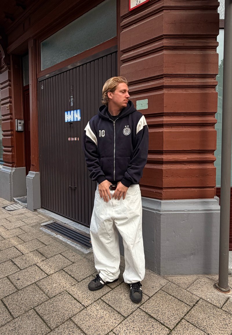 Ung mand iført sort hættejakke og hvide rummelige bukser står på fortovet ved hjørnet af en rød murstensbygning og kigger til venstre.