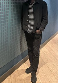 Άτομο που φοράει μαύρο μπουφάν, μαύρο παντελόνι, ριγέ πουκάμισο και μαύρα παπούτσια τύπου slip-on, στέκεται με το ένα χέρι στην τσέπη μπροστά σε μπλε τοίχο και δάπεδο από ανοιχτό ξύλο.