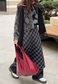 Γυναίκα που φοράει φόρεμα με μαύρο και γκρι μοτίβο, πλεκτό σακάκι μαύρο και λευκό, μαύρο μαντήλι, λευκά παπούτσια με μαύρες βούλες, κρατώντας μεγάλη κόκκινη τσάντα.