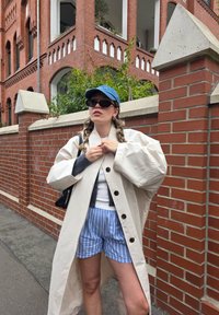 Lang beige frakk med overdimensjonerte ermer og svarte knapper lagdelt over en grå topp, parret med blåstripete shorts og en denimcap.