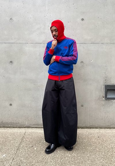 Hombre con una capucha roja y una chaqueta azul de Adidas con franjas rojas, pantalones negros de pierna ancha y zapatos negros, de pie contra una pared de cemento.