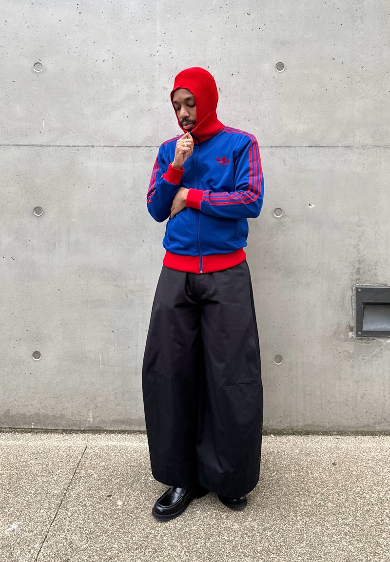 Uomo con cappuccio rosso e giacca Adidas blu con strisce rosse, pantaloni neri a gamba larga e scarpe nere, in piedi contro un muro di cemento.