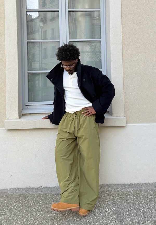 Man in black jacket, white shirt, and loose olive trousers standing against beige wall with window, looking down with hand on hip.