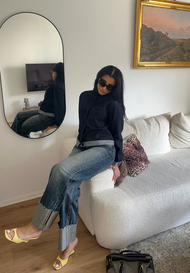 Femme portant des lunettes de soleil, une veste noire et un jean retroussé, assise sur un canapé blanc avec un coussin à motif léopard dans une pièce lumineuse avec un miroir et une peinture de paysage.